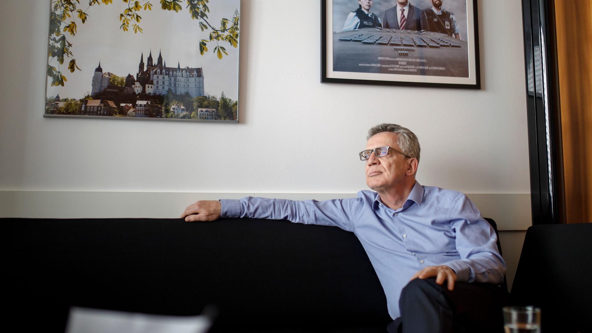 Thomas de Maiziere (CDU) sitzt entspannt auf der Couch, im Hintergrund sind einige Bilder zu erkennen. 