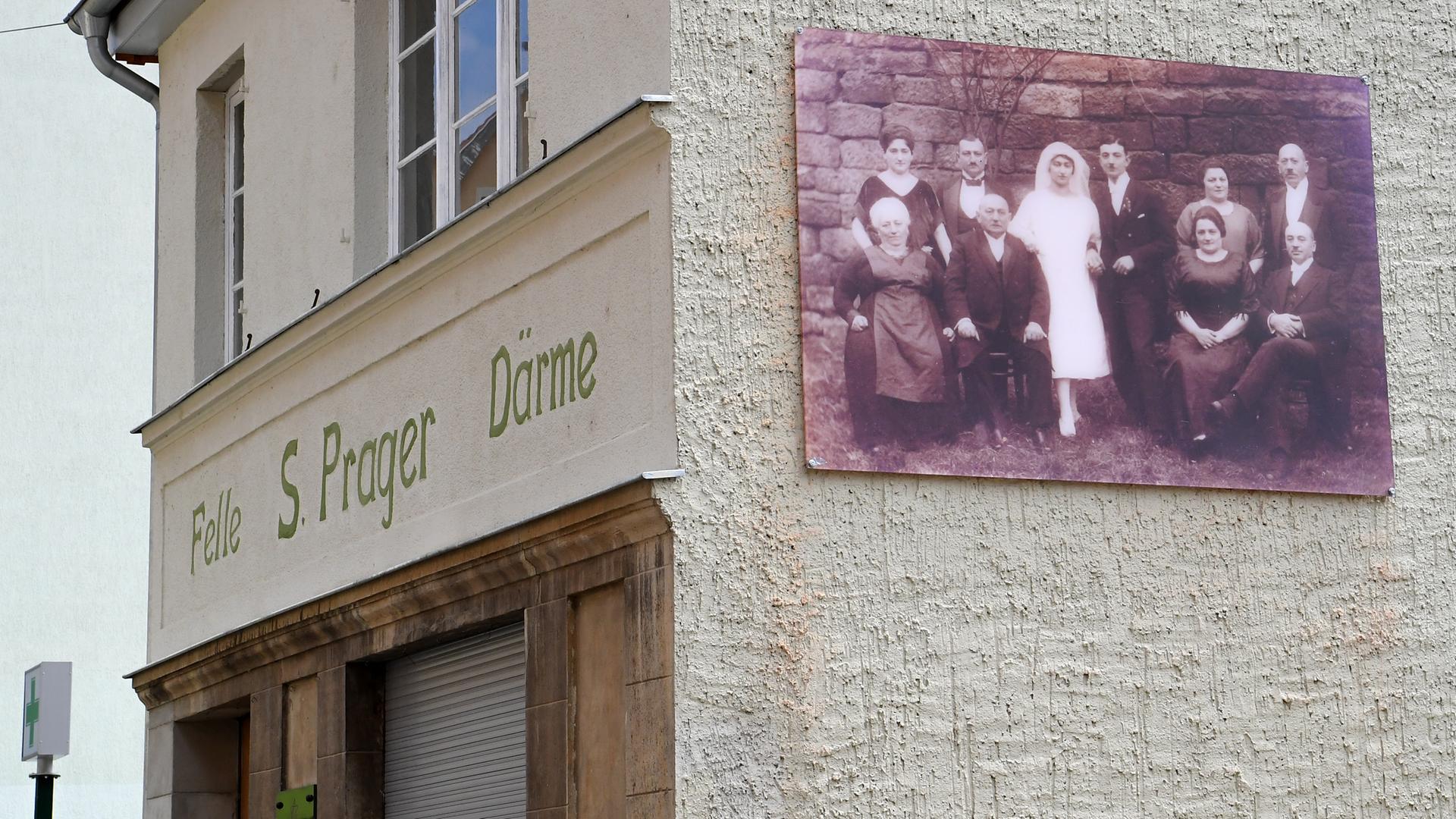 Blick auf die Front und eine Seite des kleinen alleinstehenden Hauses. Auf der Front steht "Felle - S. Prager - Därme". Auf der Seite ist das große historische Bild der Familie angebracht.