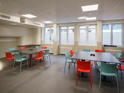Blick in einen Raum mit großen Tischen mit mehreren Stühlen. An der Decke hängen Entlüftungsanlagen.