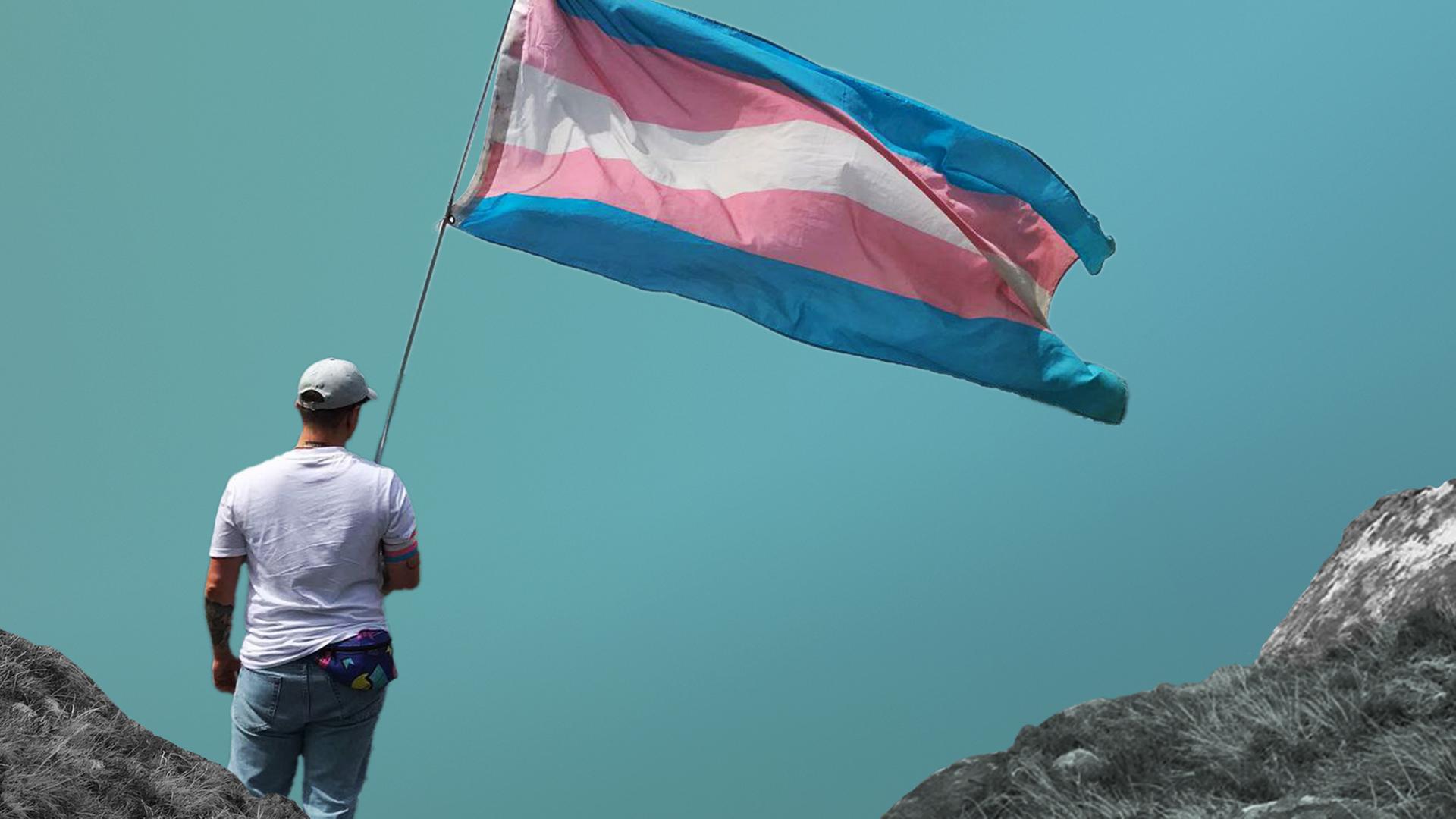 Dean steht in Jeans und weißem T-Shirt von hinten fotografiert mit einer blau-rosa-weiß gestreiften Transflagge in der Hand vor einem blauen Hintergrund mit Bergen.