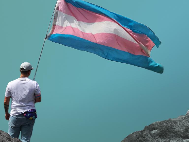 Dean steht in Jeans und weißem T-Shirt von hinten fotografiert mit einer blau-rosa-weiß gestreiften Transflagge in der Hand vor einem blauen Hintergrund mit Bergen.
