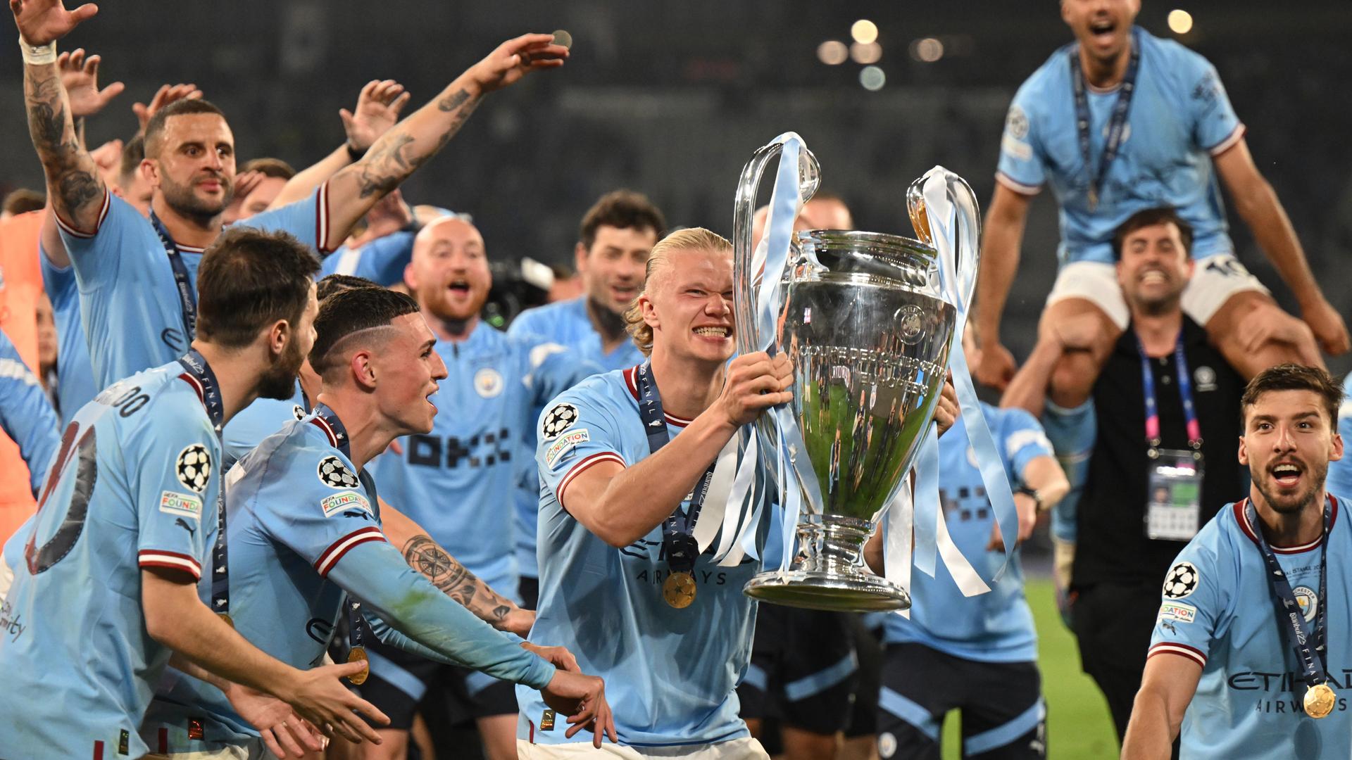 Manchester Citys Spieler Erling Haaland hält den Champions-League-Pokal in den Händen, neben ihm stehen jubelnde Mitspieler. Manchester Citys Spieler Erling Haaland hält den Champions-League-Pokal in den Händen, neben ihm stehen jubelnde Mitspieler.