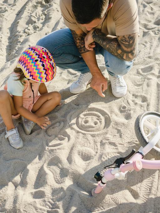 Vater und Tochter sitzen am Strand und malen lachende Gesichter in den Sand.