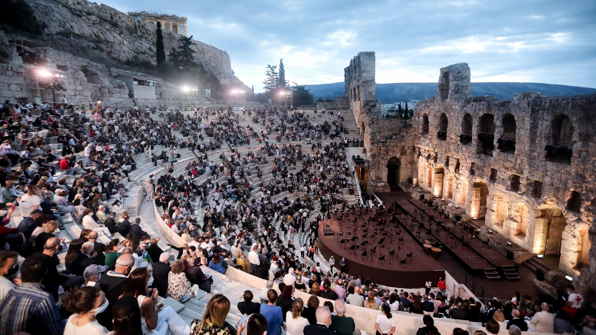 Bei Dämmerung sitzen Zuschauer im Freien im Odeion-Theater in Athen. Auf der Bühne sind Stühle für ein Orchester.