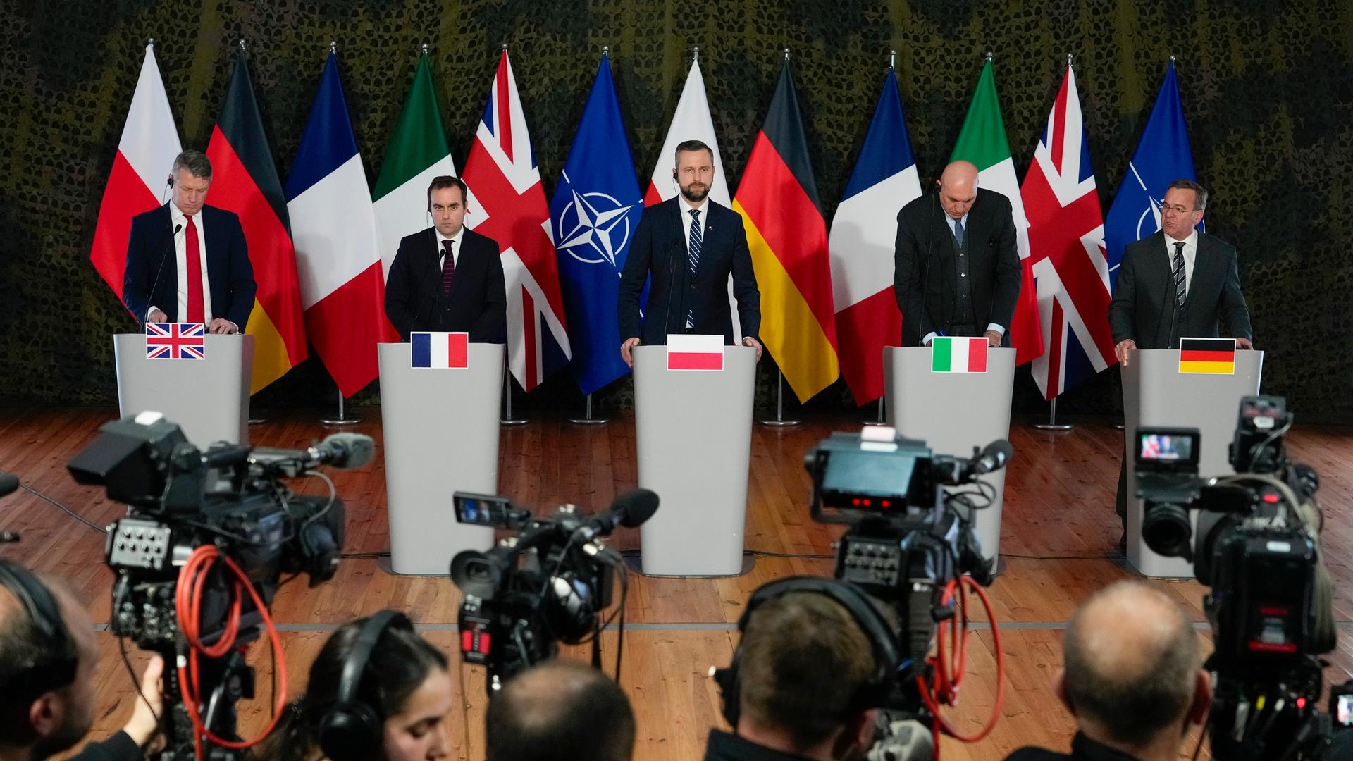 Beim Treffen der Fünfergruppe in Warschau stehen die Verteidigungsminister bzw. Vertreter aus Großbritannien, Frankreich, Polen, Italien und Deutschland nebeneinander hinter Rednerpulten. Im Hintergrund stehen viele Fahnen. 