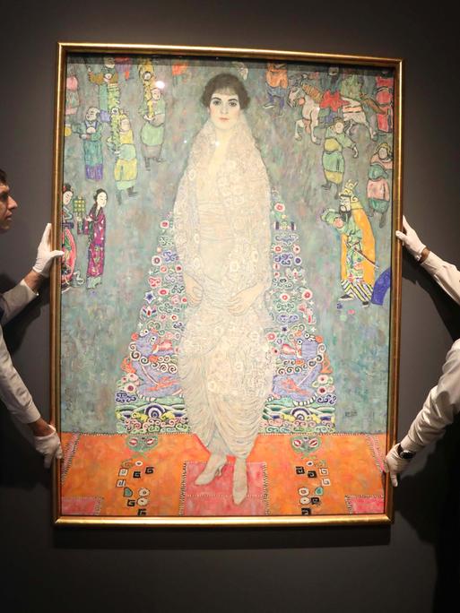 Zwei Männer halten ein Gemälde von Gustav Klimt, sie tragen Schürze und weiße Handschuhe