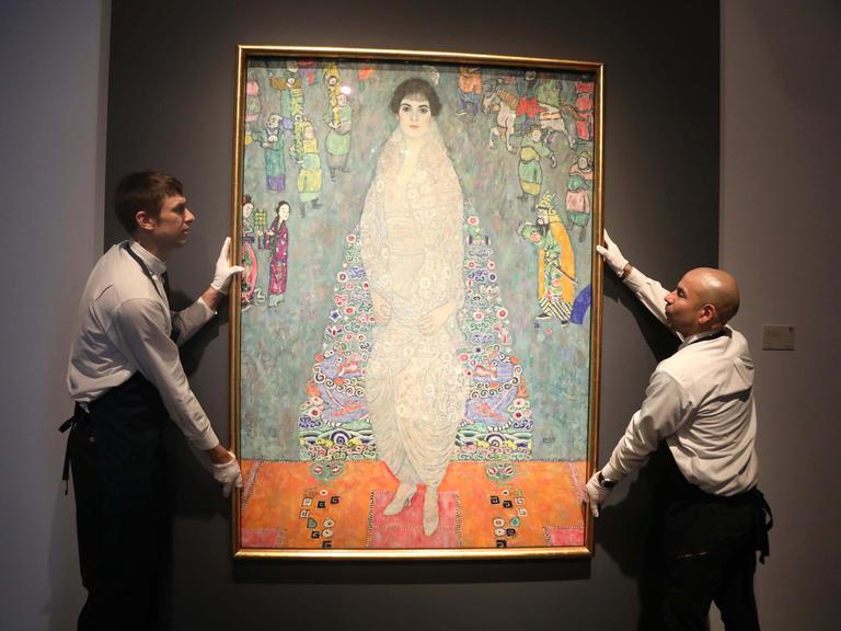 Zwei Männer halten ein Gemälde von Gustav Klimt, sie tragen Schürze und weiße Handschuhe