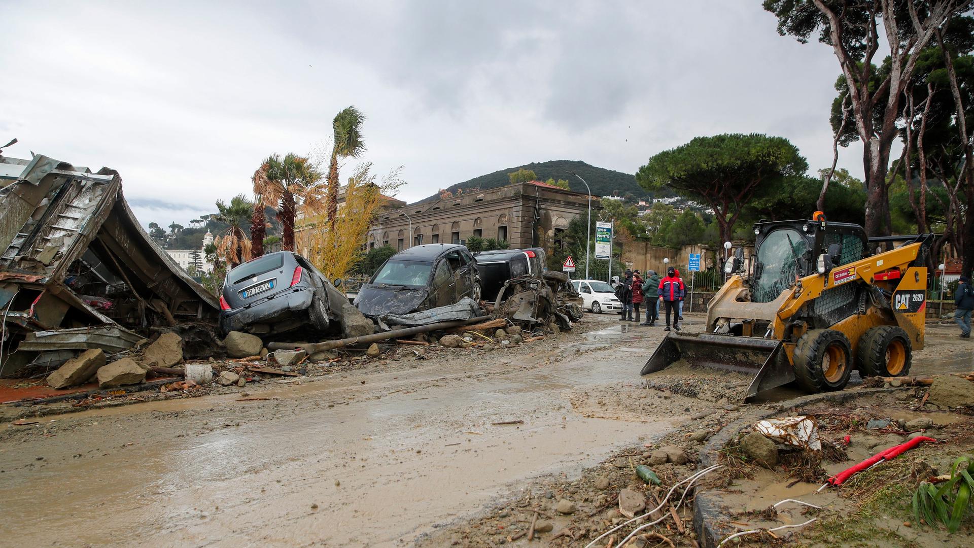 Eine Straße im Schlamm, an deren Rändern Räumfahrzeuge, kaputte Autos und ein eingestürztes Gebäude.