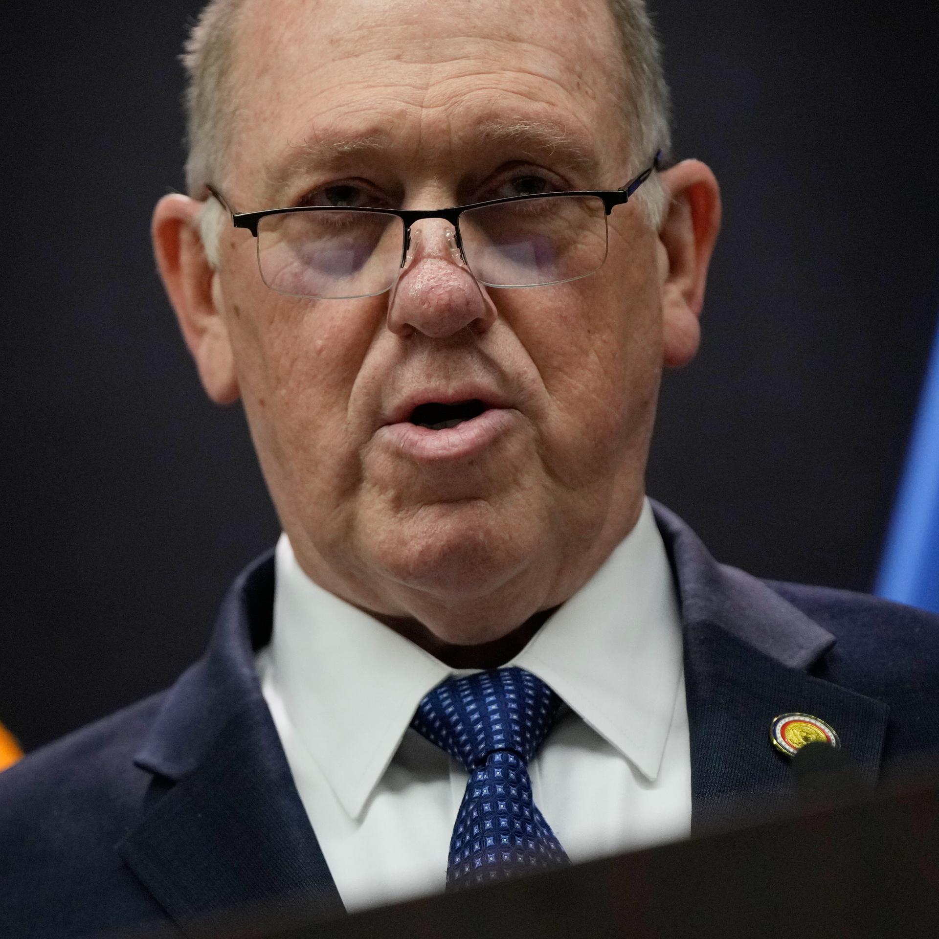 Tom Homan schaut bei einer Pressekonferenz in Minneapolis in die Kamera. Er trägt ein weißes Hemd, eine Anzugjacke und eine blaue Krawatte.