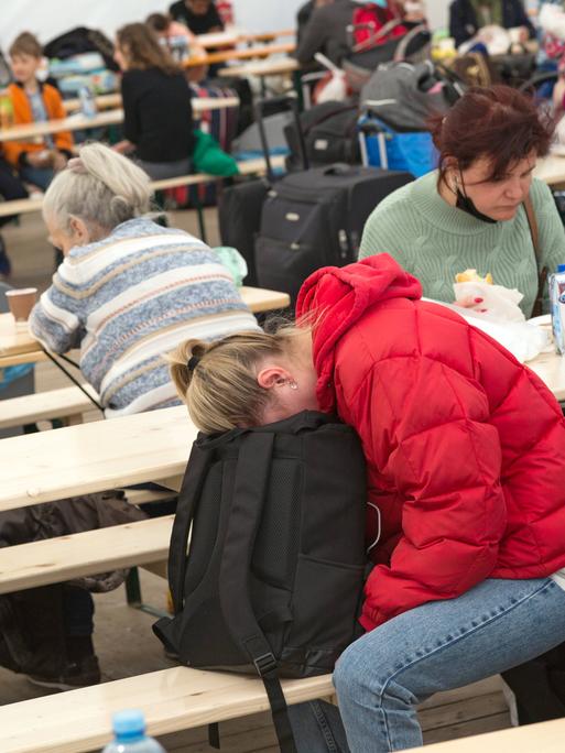 Erschöpfte ukrainische Flüchtlinge sitzen im Willkommenszelt am Berliner Hauptbahnhof auf Holzbänken. 