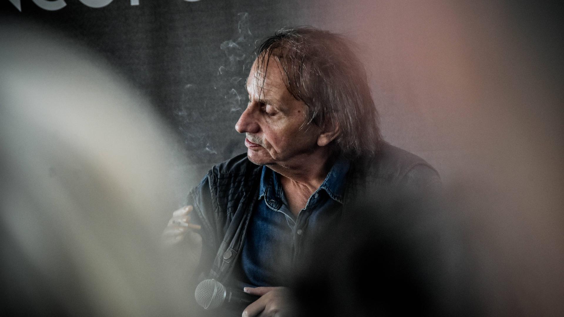 Michel Houellebecq sitzt mit genervtem Gesichtsausdruck auf einem Podium