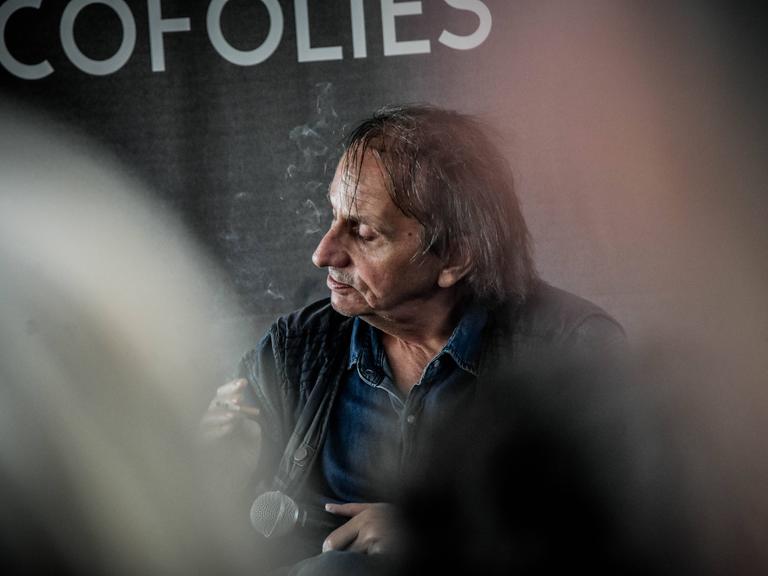 Michel Houellebecq sitzt mit genervtem Gesichtsausdruck auf einem Podium