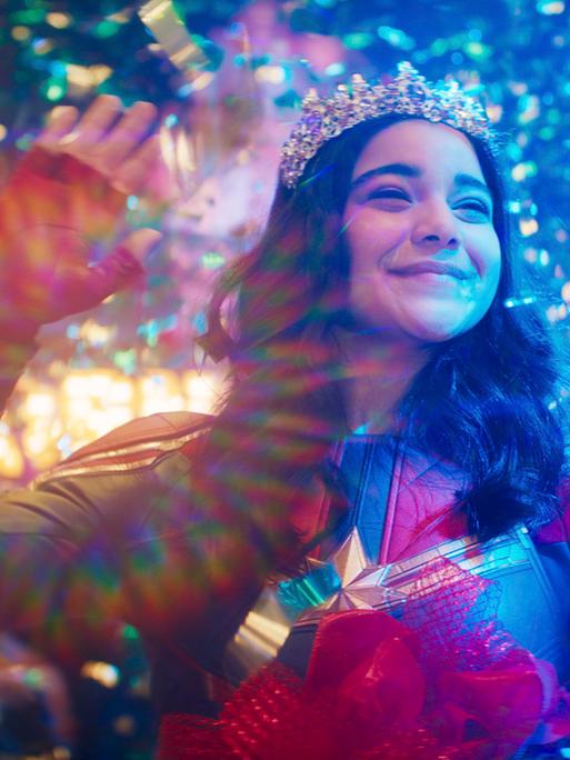 Im Still aus "Ms. Marvel" steht ein Mädchen mit einem Diadem auf dem Kopf winkend in einem Regen aus buntem Glitzerkonfetti.