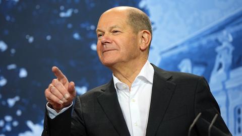 Das Foto zeigt Bundeskanzler Scholz im schwarzen Jackett. Scholz steht vor einem  Bild von Schloss Meseberg, lächelt und zeigt mit dem rechten Zeigefinger nach vorne.