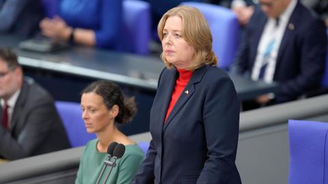 Bas im schwarzen Jackett steht an ihrem Platz und schaut ernst. Dahinter unscharf weitere personen auf der Regierungsbank.