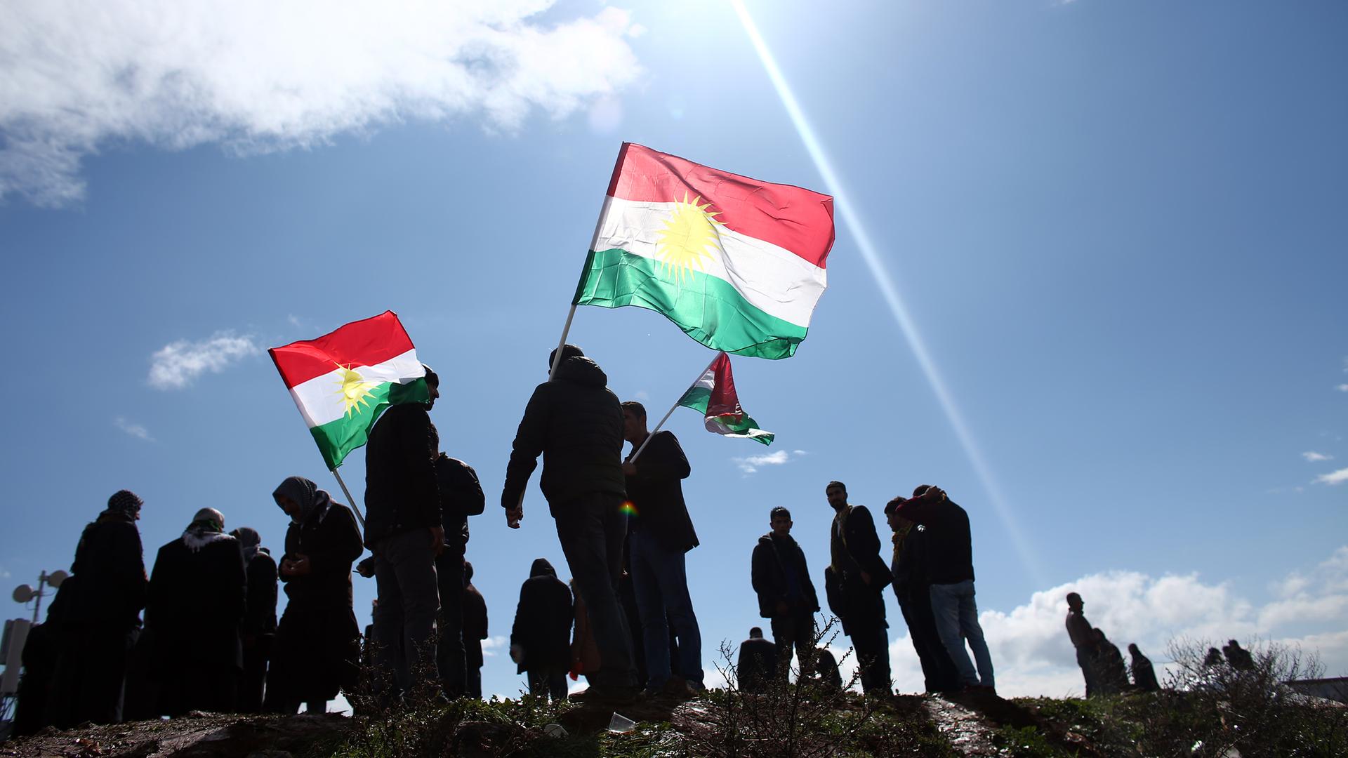 Eine Gruppe von Menschen schwennken vor strahlend blauem Himmel die kurische Flagge mir ihren Farben rot, weiß, grün und einem Sonnensymbol in der Mitte.