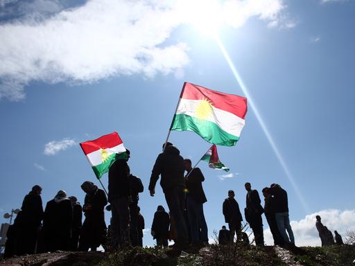 Eine Gruppe von Menschen schwennken vor strahlend blauem Himmel die kurische Flagge mir ihren Farben rot, weiß, grün und einem Sonnensymbol in der Mitte.