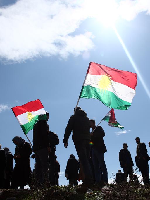 Eine Gruppe von Menschen schwennken vor strahlend blauem Himmel die kurische Flagge mir ihren Farben rot, weiß, grün und einem Sonnensymbol in der Mitte.