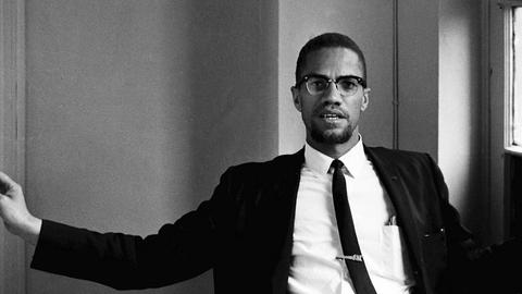 Malcolm X sitzt im Anzug und Krawatte im Foyer eines Hotels.