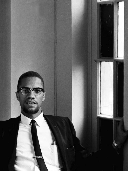 Malcolm X sitzt im Anzug und Krawatte im Foyer eines Hotels.