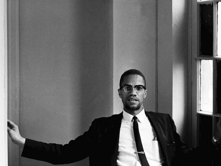 Malcolm X sitzt im Anzug und Krawatte im Foyer eines Hotels.