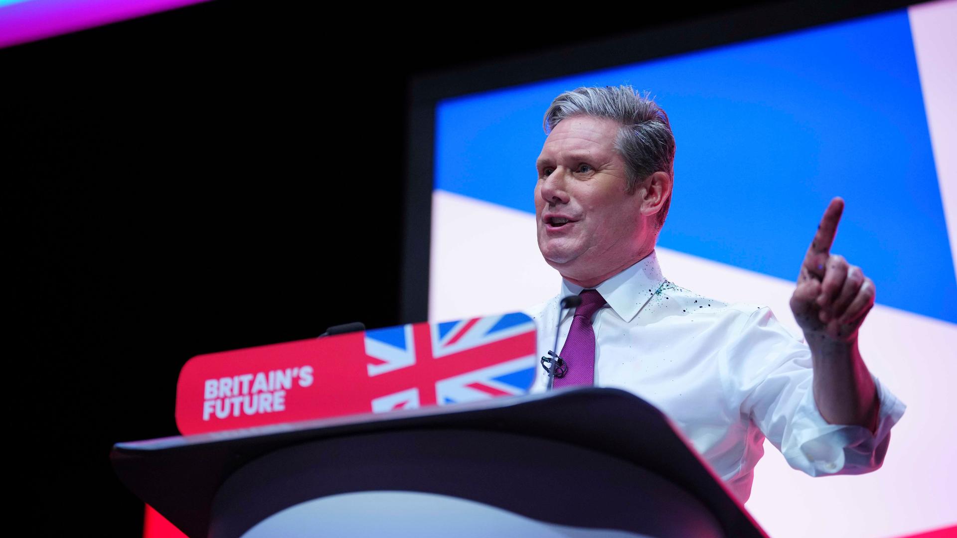 Starmer steht am Rednerpult mit dem Union Jack, spricht in ein Mikrofon und zeigt mit dem linken Zeigefinger nach oben.