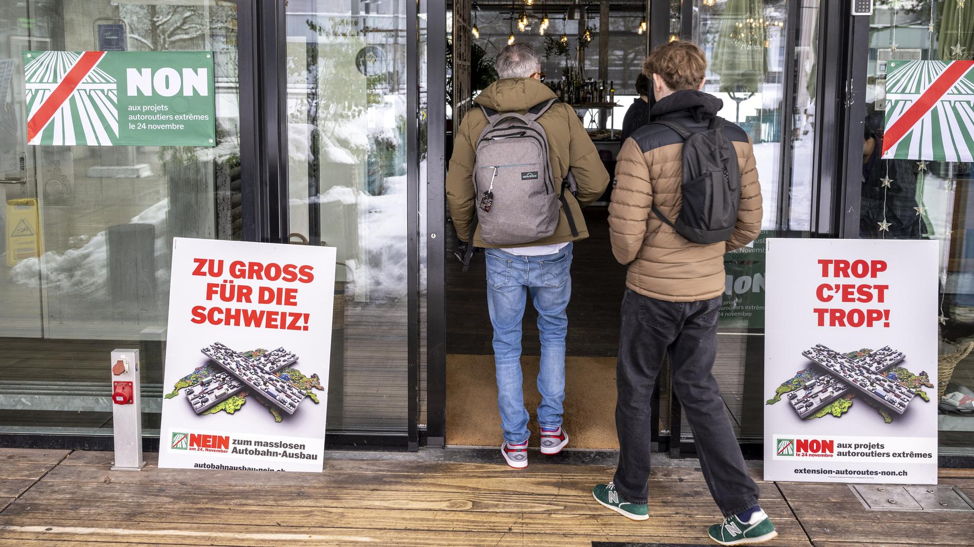 Zwei Männer in Winterjacken und Rucksäcken betreten ein Stimmlokal mit einer gläsernen Front. Davor und an den Fenstern Plakete gegen den Autobahnausbau.