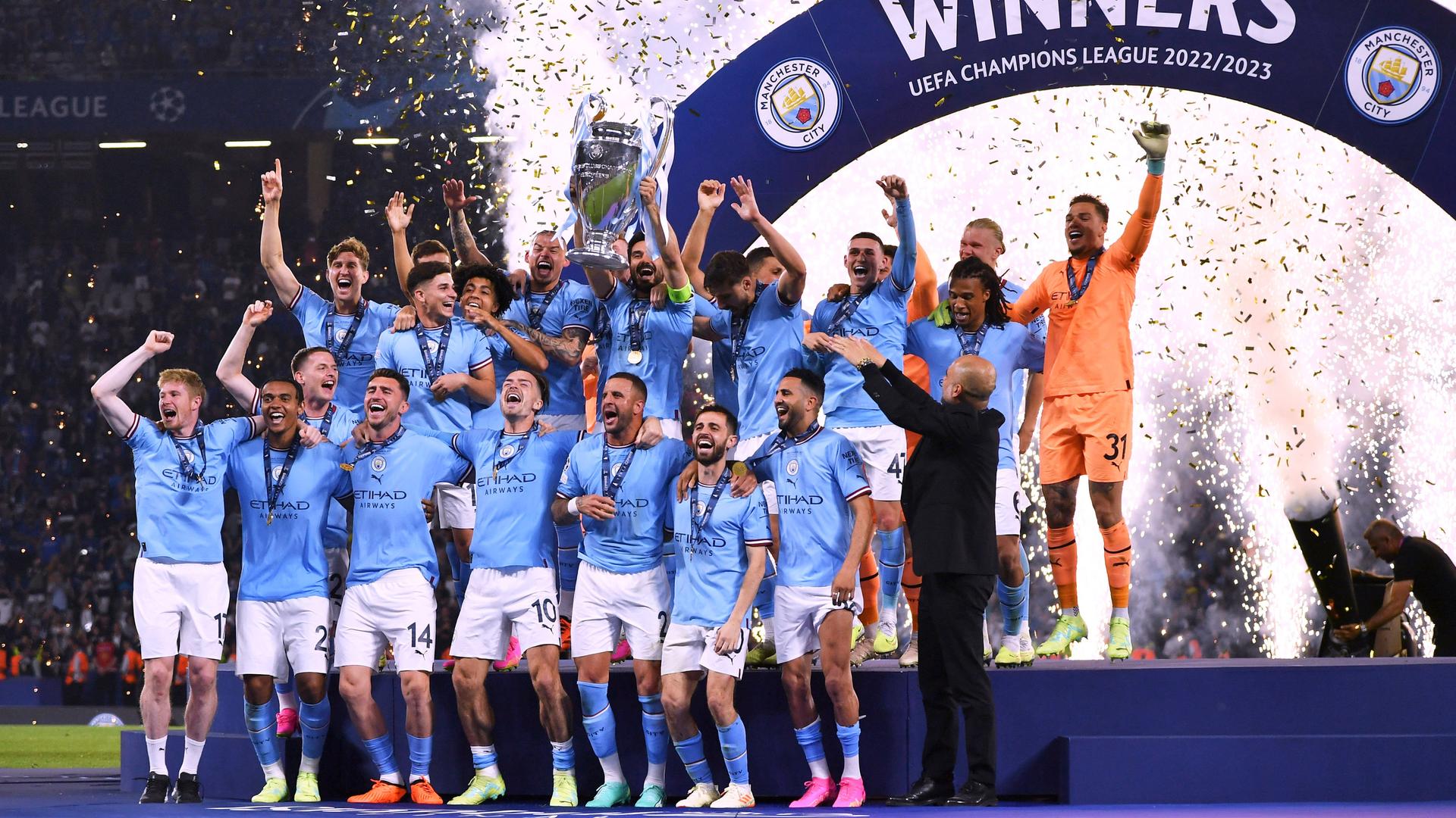 Die Spieler von Manchester City feiern den Sieg beim Champions League-Finale auf einer Tribüne. Ein Spieler streckt den großen Pokal in die Luft. Im Hinter-Grund ist Feuer-Werk und Konfetti zu sehen. 