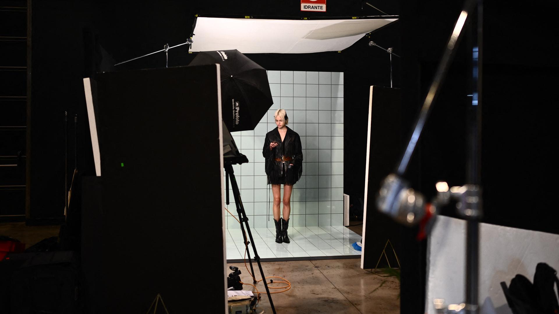 Ein blondes Model steht während einer Modenschau wartend in der Kulisse. Klein im Bild und von Equipment umgeben wirkt sie verloren.