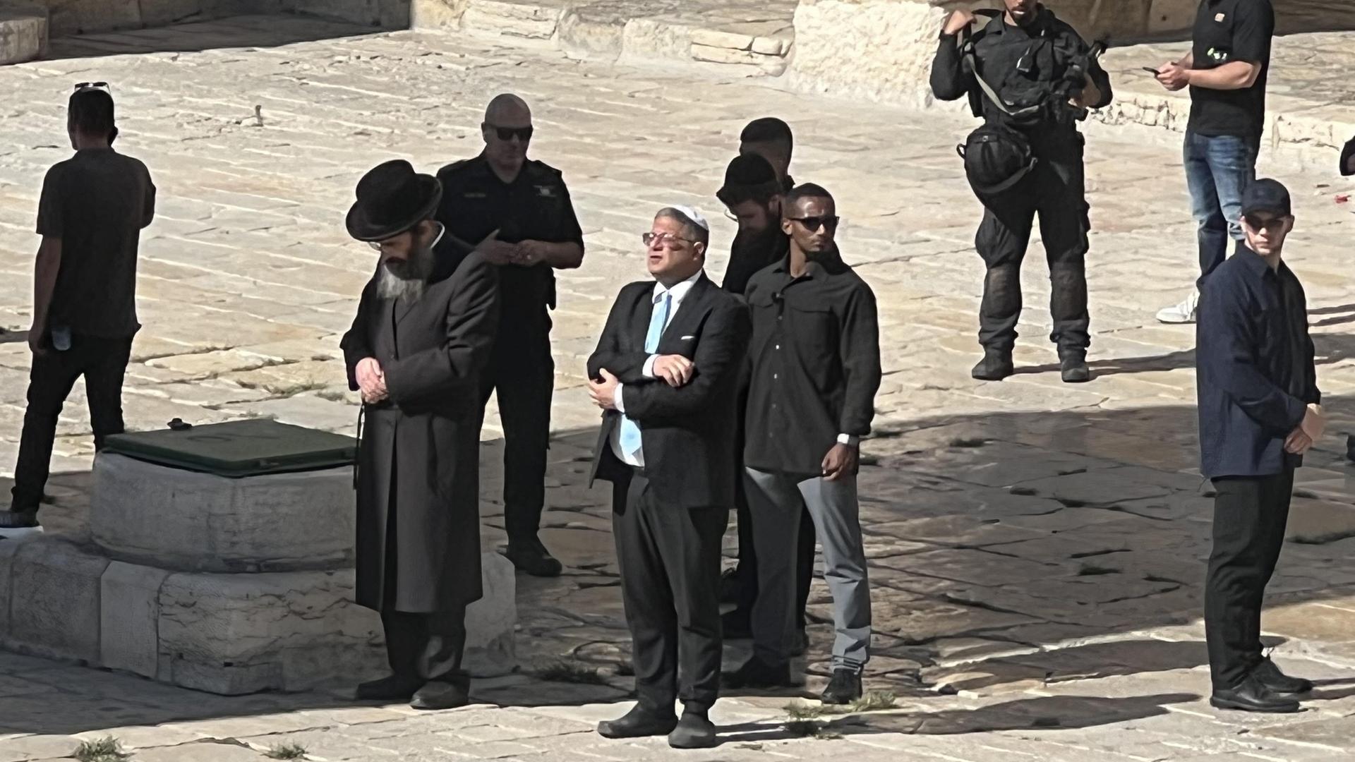 Der israelische Polizeiminister Ben-Gvir steht mit verschränkten Armen auf dem Tempelberg. Neben ihm steht ein betender Rabbi. Um die beiden herum Leibwächter und Sicherheitsleute.