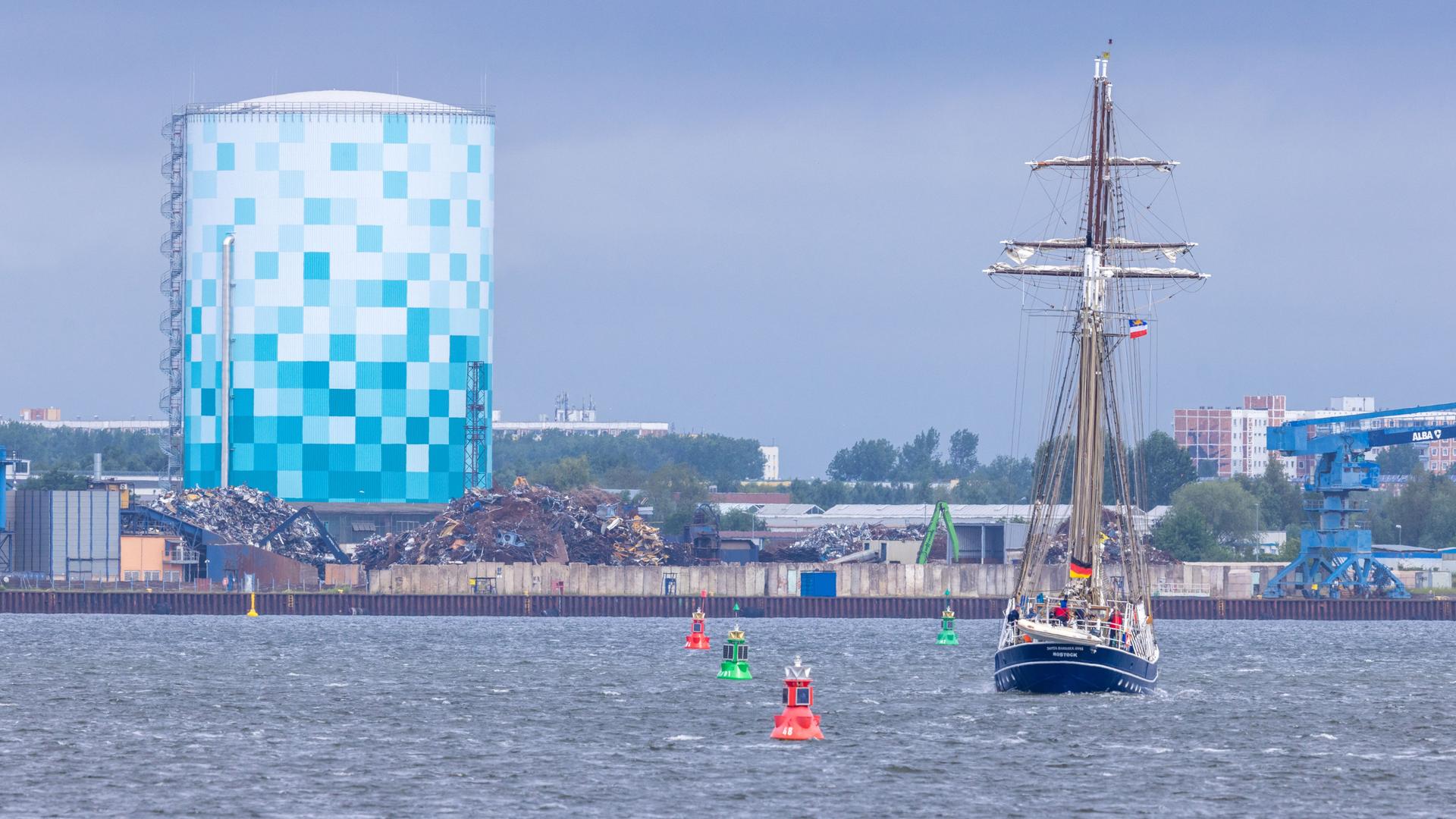 Auf dem Gelände der Stadtwerke Rostock an der Warnow steht ein druckloser Warmwasserspeicher. Der Speicher ist insgesamt 94 Meter hoch, 56 Meter sind über dem Erdboden sichtbar. Davor fährt ein Segelschiff vorbei.