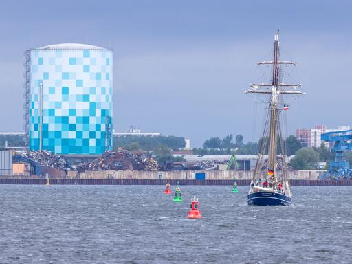 Auf dem Gelände der Stadtwerke Rostock an der Warnow steht ein druckloser Warmwasserspeicher. Der Speicher ist insgesamt 94 Meter hoch, 56 Meter sind über dem Erdboden sichtbar. Davor fährt ein Segelschiff vorbei.