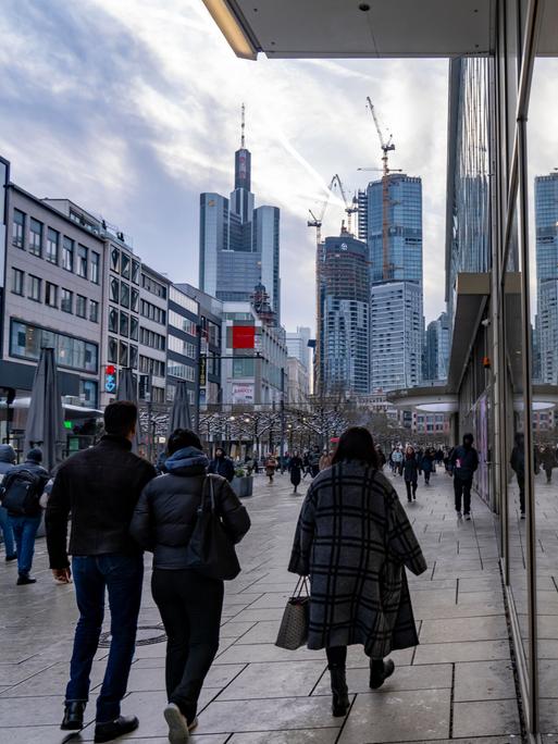 Menschen laufen durch die Einkaufsstraße Zeil in Frankfurt am Main, am Horizont ist die Skyline zu sehen.