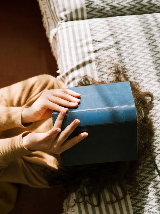 Foto einer jungen Frau, die auf dem Sofa liegt und ein Buch vor ihr Gesicht hält. Foto einer jungen Frau, die auf dem Sofa liegt und ein Buch vor ihr Gesicht hält.