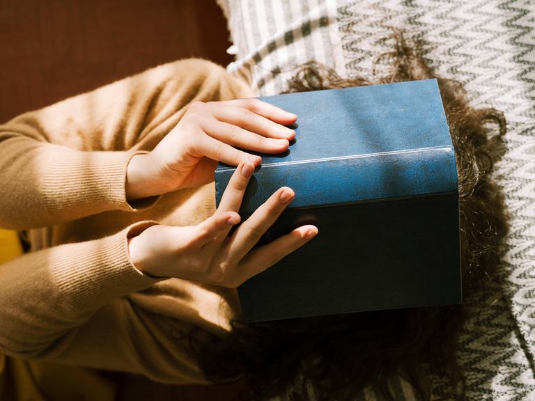 Foto einer jungen Frau, die auf dem Sofa liegt und ein Buch vor ihr Gesicht hält. Foto einer jungen Frau, die auf dem Sofa liegt und ein Buch vor ihr Gesicht hält.