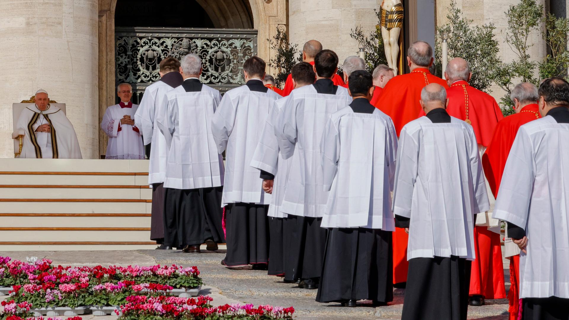 Die Kardinäle gehen in einer Doppelreihe die Stufen zum Papst hoch; man sieht sie von hinten. Am linken Bildrand sieht man Franziskus in seinem Stuhl sitzen. Am rechten Bildrand ein großes Kruzifix mit dem blutenden Jesus.