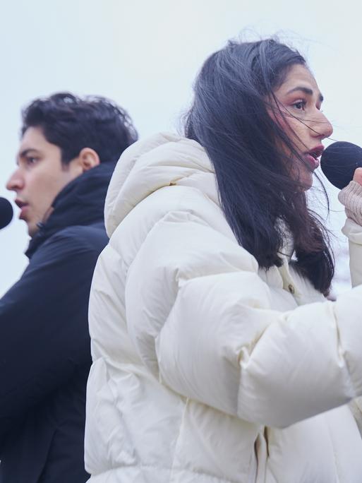 Der Rapper Apsilon und die Rapperin Wa22ermann stehen Rücken an Rücken auf einer Bühne. Sie tragen dicke Winterjacken und halten Mirkofone in ihren Händen.