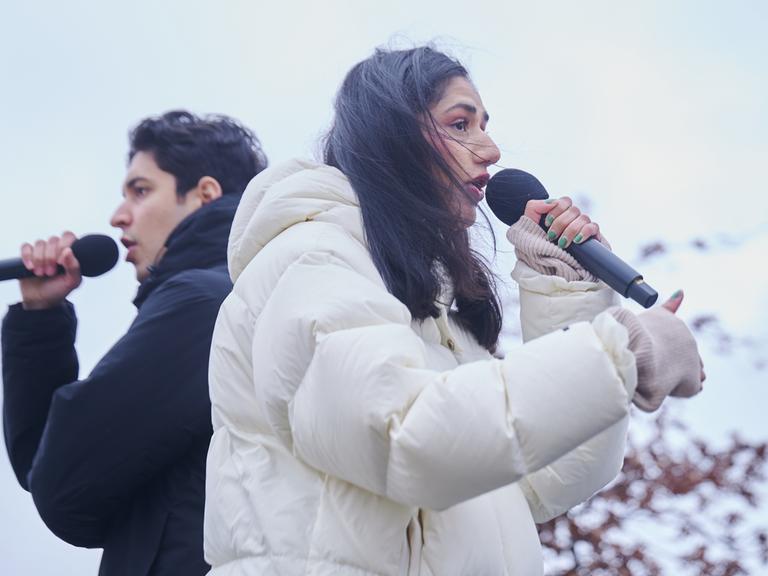 Der Rapper Apsilon und die Rapperin Wa22ermann stehen Rücken an Rücken auf einer Bühne. Sie tragen dicke Winterjacken und halten Mirkofone in ihren Händen.