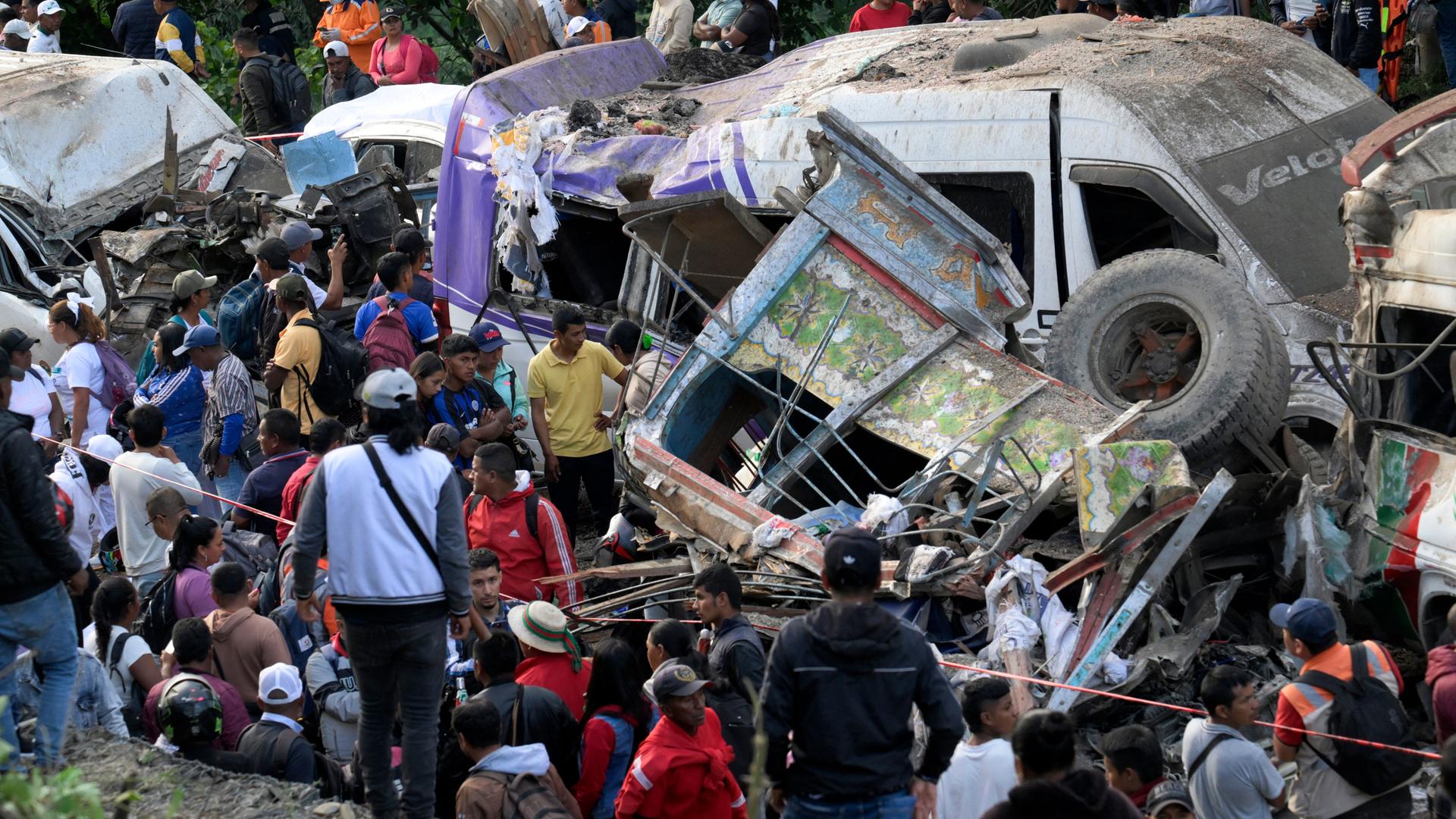 Das Bild zeigt mehrere zerstörte und schwer beschädigte Fahrzeuge. Rund um die Trümmer stehen viele Menschen.