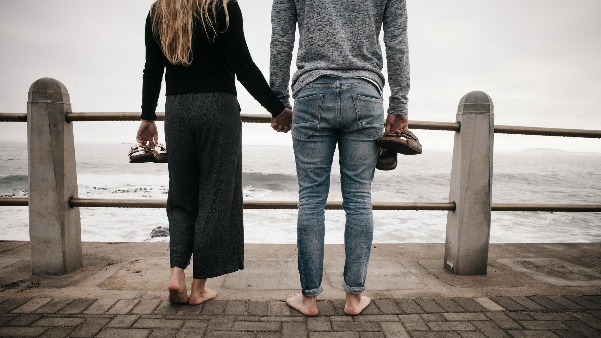 Ein Paar steht baarfuß und Händchen haltend an einem Geländer. Dahinter beginnt das Meer, der Himmel ist bedeckt und grau.