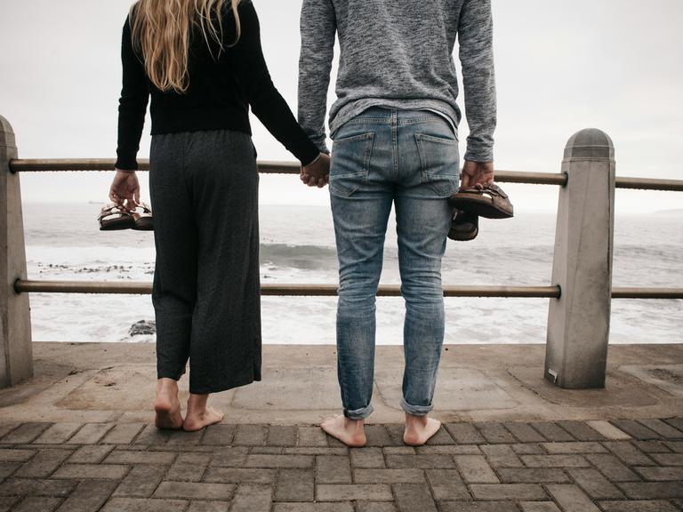 Ein Paar steht baarfuß und Händchen haltend an einem Geländer. Dahinter beginnt das Meer, der Himmel ist bedeckt und grau.