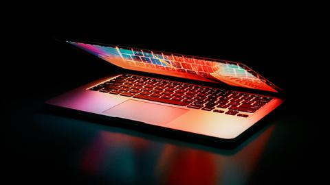 Ein Laptop mit butem Display liegt beinahe zusammengeklappt in einem dunklen Raum, sodass bunte Farben aus dem Laptop strahlen.