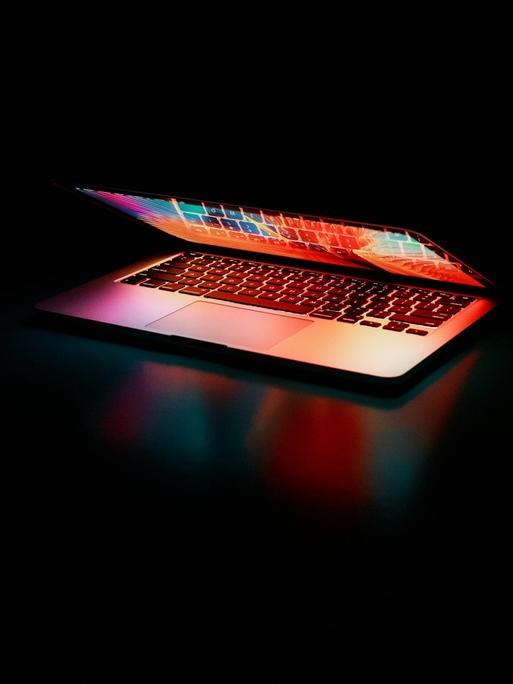 Ein Laptop mit butem Display liegt beinahe zusammengeklappt in einem dunklen Raum, sodass bunte Farben aus dem Laptop strahlen.