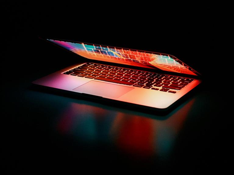 Ein Laptop mit butem Display liegt beinahe zusammengeklappt in einem dunklen Raum, sodass bunte Farben aus dem Laptop strahlen.