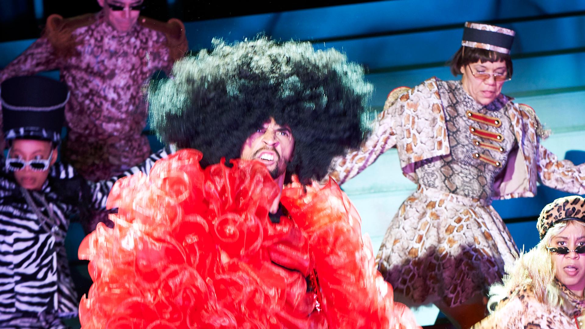 Schauspieler tragen auf einer Theaterbühne Kostüme, die an Karneval erinnern.