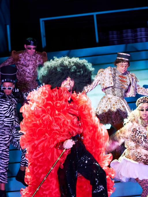 Schauspieler tragen auf einer Theaterbühne Kostüme, die an Karneval erinnern.
