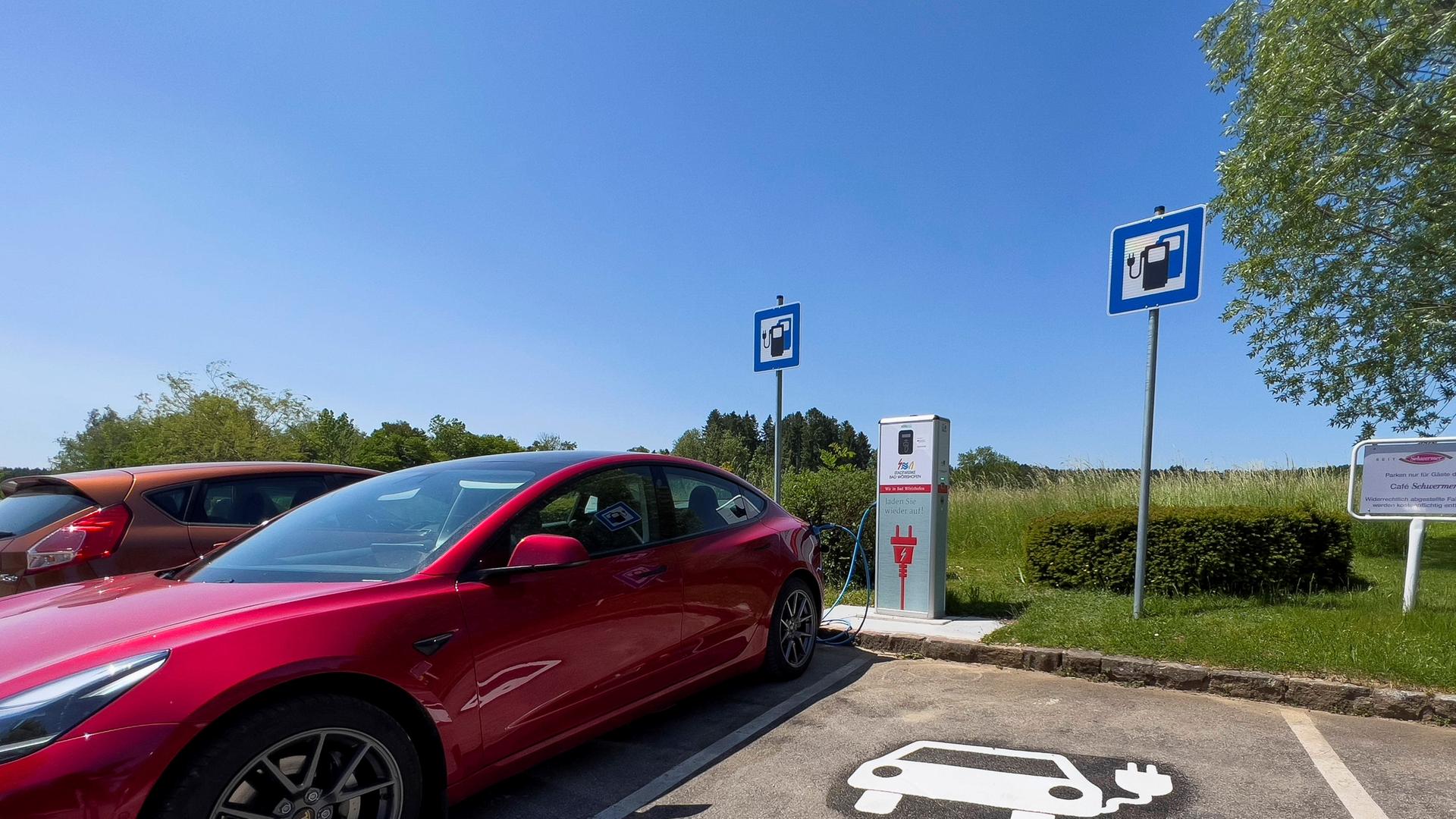 Ein roter Tesla Model 3 lädt seinen Akku an einer Ladesäule voll auf einem Parkplatz, mit Wiese und Bäumen daneben (08.06.2023) 