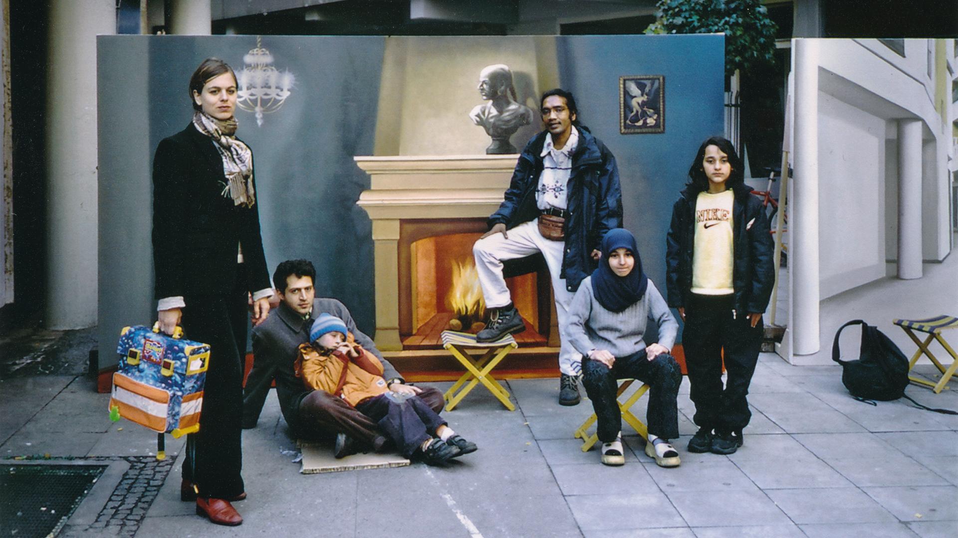 Ein Gruppe von Menschen posiert vor einem Gemälde. Das Bild zeigt ein Zimmer mit einem brennenden Kamin.