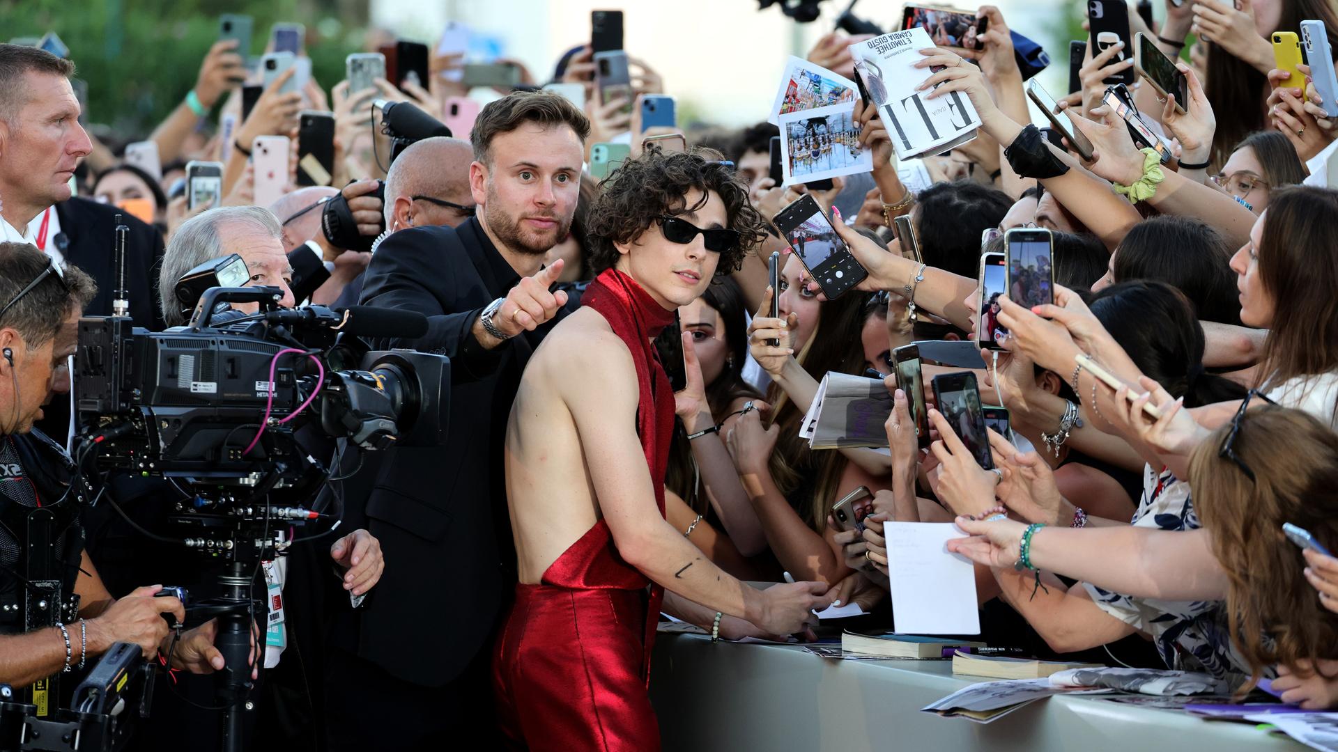 Timothée Chalamet steht vor einer Absperrung und signiert Autogramme, während auf der anderen Seite des Zauns dutzende Fans begeistert ihre Smartphones auf ihn richten.