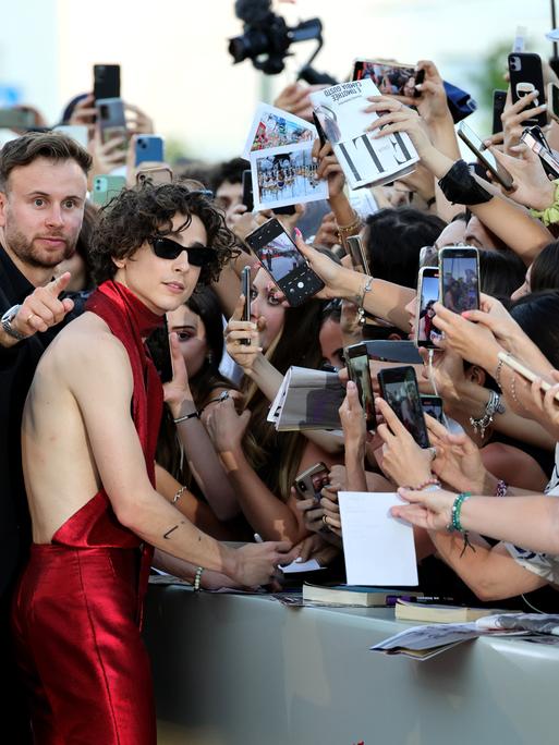 Timothée Chalamet steht vor einer Absperrung und signiert Autogramme, während auf der anderen Seite des Zauns dutzende Fans begeistert ihre Smartphones auf ihn richten.